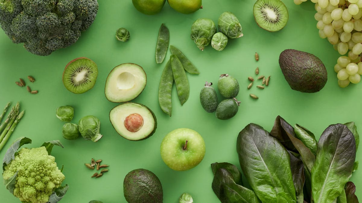 Top view of assorted fresh green vegetables and fruits on a green background, perfect for healthy lifestyle visuals.